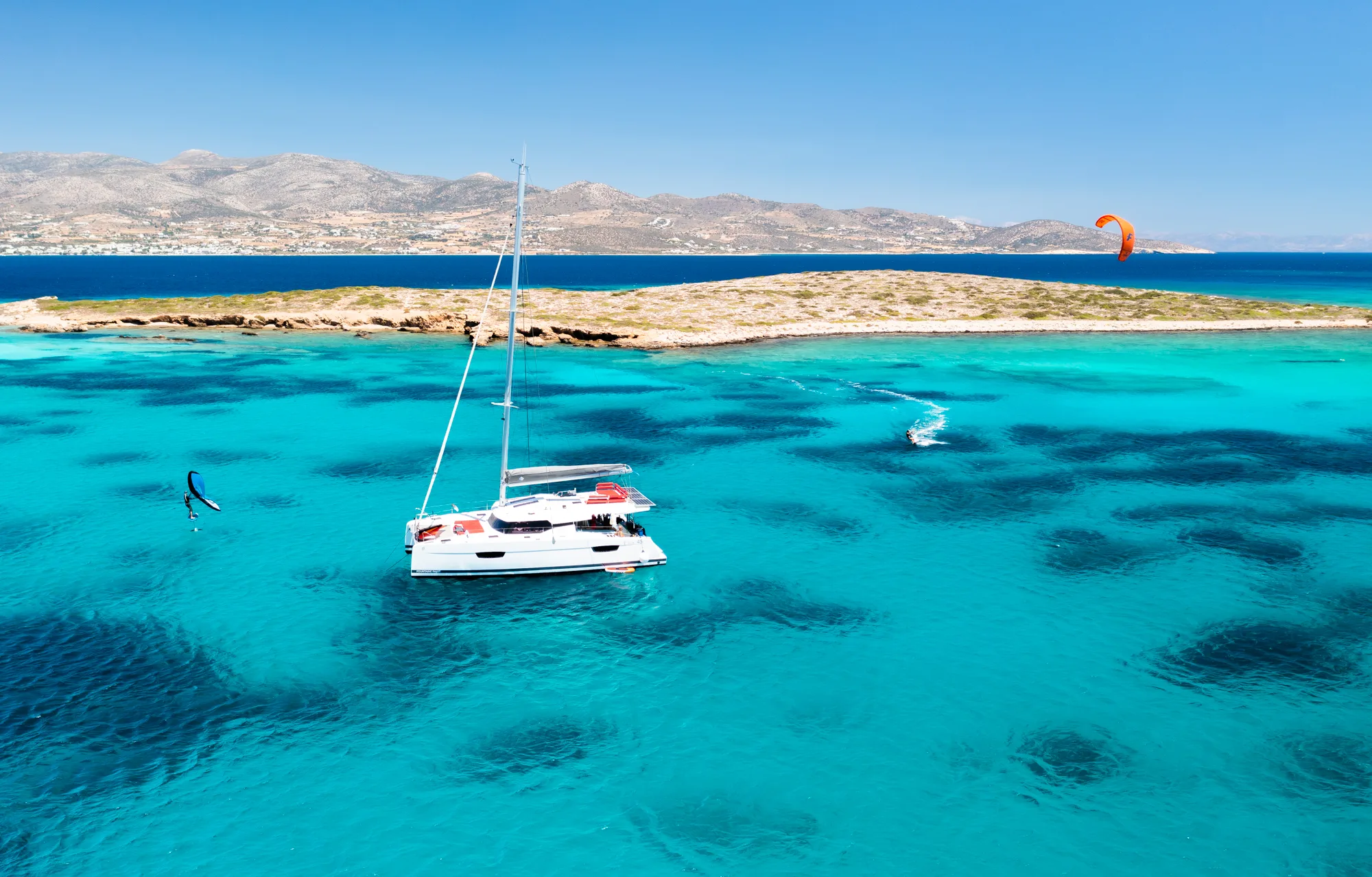Aerial view of Makani anchored off a remote island in turquoise water, with a kitesurfer and wing foiler riding nearby.