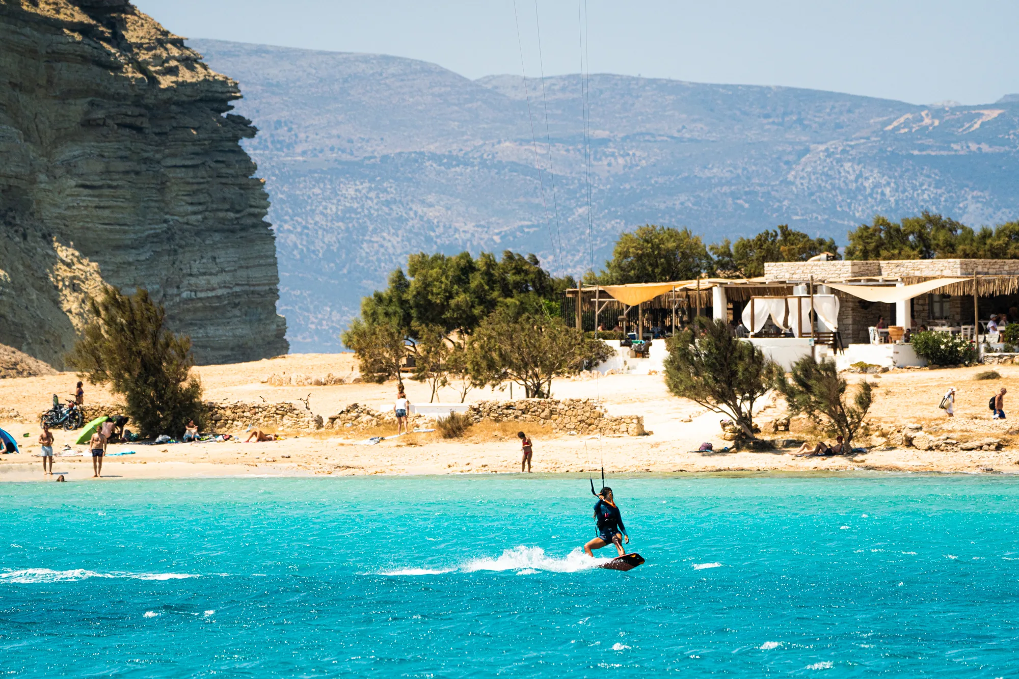Kitesurfer cruising shallow turquoise water in front of a Cycladic beach bar.