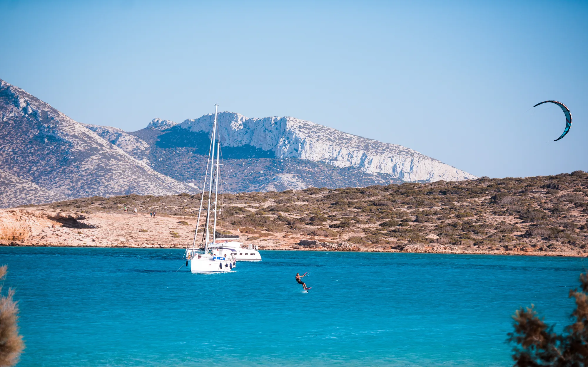 Kitesurfer riding turquoise water beside the Makani catamaran anchored off a Cyclades island.