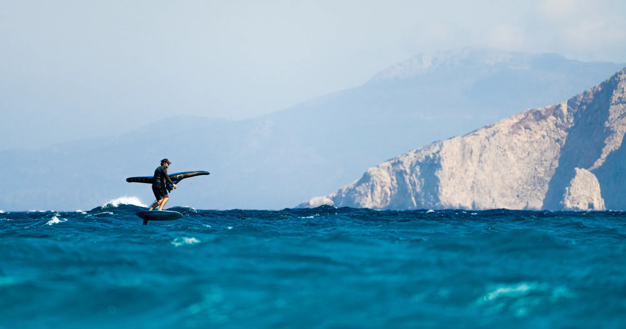 A wing foiler carving open water with dramatic Cycladic cliffs behind.